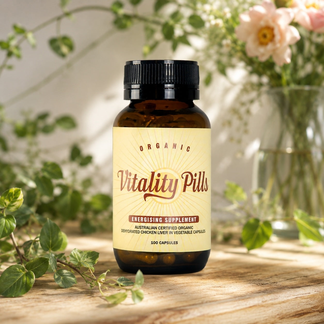 An amber glass bottle of Vitality Pills dietary supplement with a cream label and black cap, placed on a wooden surface with blurred green leaves and pink flowers in the background.