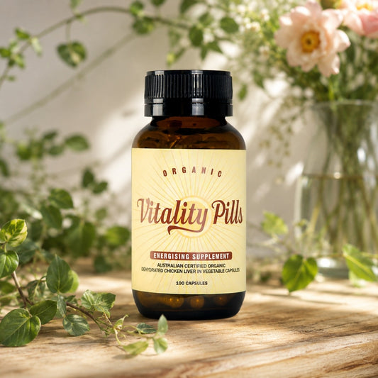 An amber glass bottle of Vitality Pills dietary supplement with a cream label and black cap, placed on a wooden surface with blurred green leaves and pink flowers in the background.